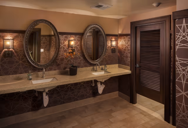 Interior view of a bathroom with two oval mirrors above a long beige countertop with two sinks. The wall behind the sinks has a decorative geometric pattern and two wall-mounted lights. There is a dark wooden door to the right.
