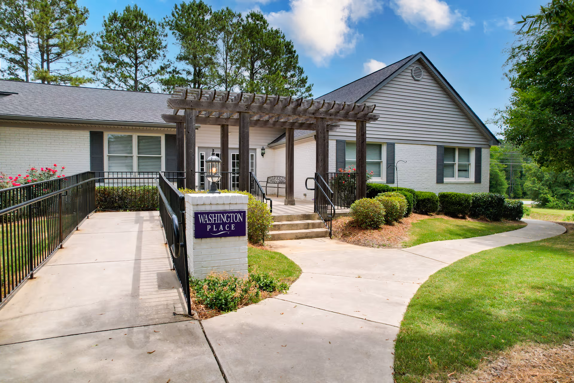 Exterior view of a single-story building with white brick walls and gray siding. There is a concrete walkway with a ramp and stairs leading to the entrance, which is covered by a wooden pergola. The area is landscaped with bushes, grass, and trees under a partly cloudy sky. A sign near the entrance reads 'Washington Place'.