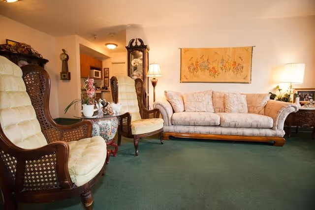 A cozy living room with a green carpet, a light-colored floral sofa, two wooden armchairs with cushioned seats, a small round table with plants and decorative items, a tall grandfather clock, a floor lamp, and a framed floral artwork hanging on the wall.