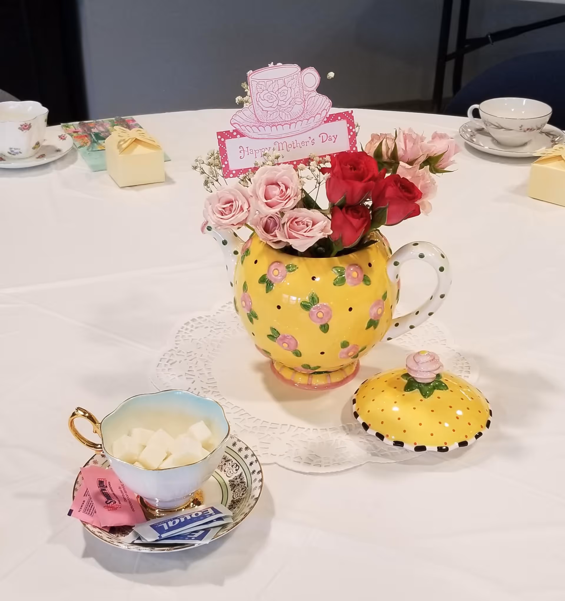 A yellow polka-dotted teapot used as a vase with pink and red roses and a 'Happy Mother's Day' card on a white tablecloth, surrounded by teacups, saucers, sugar cubes and tea packets.