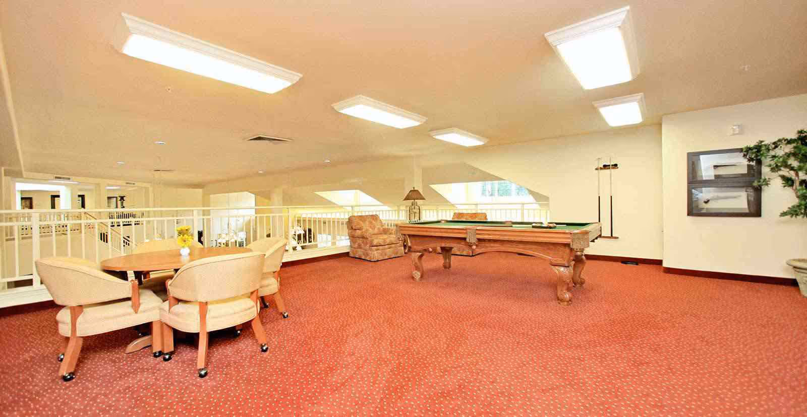 A spacious recreational room with red carpet flooring featuring a wooden pool table, a round table with four beige chairs, a cushioned armchair, a lamp on a small table, framed artwork on the wall, and a potted plant in the corner. The room has white walls, large ceiling lights, and windows letting in natural light.