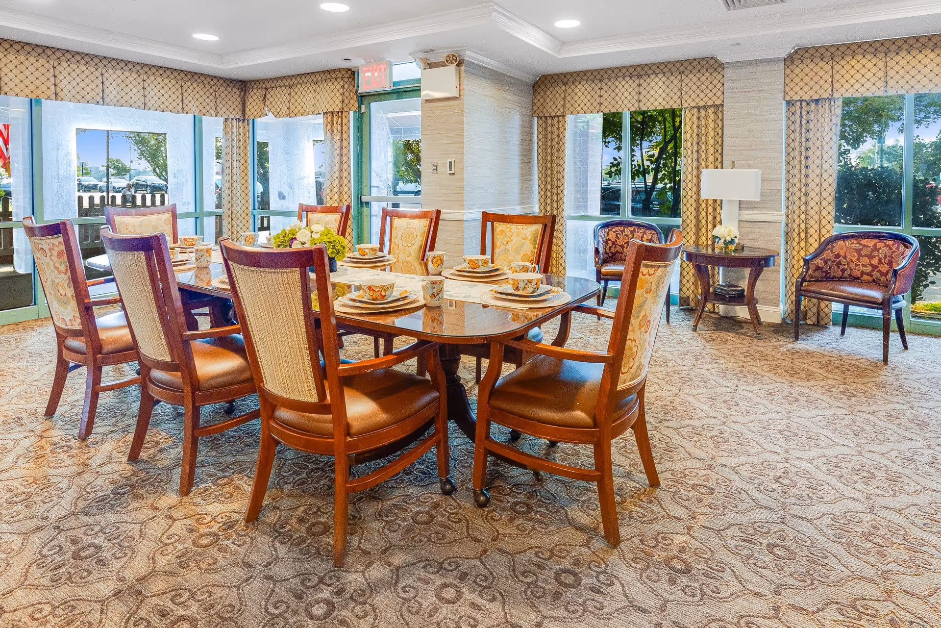 A well-lit dining room with a large wooden dining table set for eight people with matching chairs. The table is arranged with plates, bowls, cups, and a centerpiece of flowers. The room has large windows with patterned curtains, allowing natural light to enter. There are two upholstered armchairs and a small round table with a lamp in the corner. The carpet has a decorative pattern, and an exit door is visible in the background.