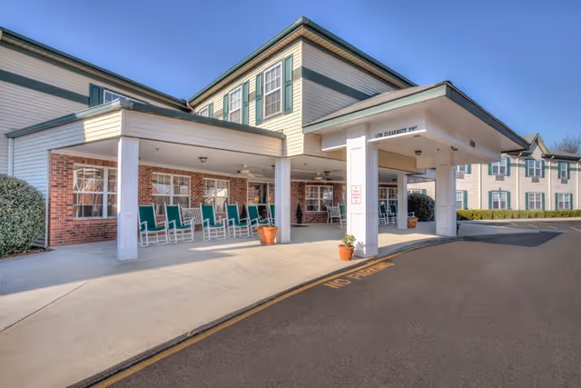 Covered front entrance of a senior living building with a row of green rocking chairs on the porch and potted plants by the drive-up canopy.