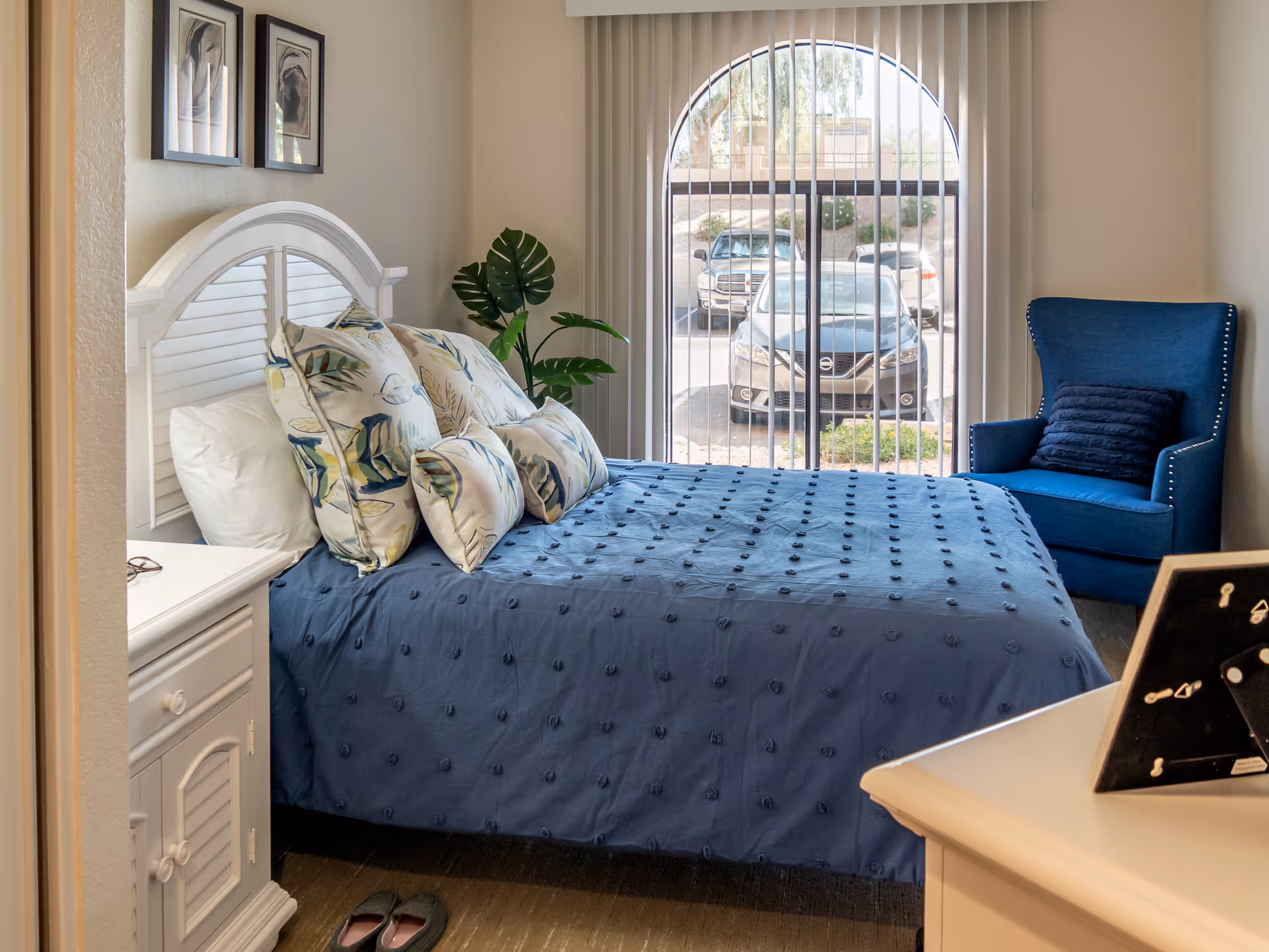Sunlit bedroom with a blue quilted bed and patterned pillows, white headboard, a blue armchair, and an arched window with vertical blinds showing parked cars outside.