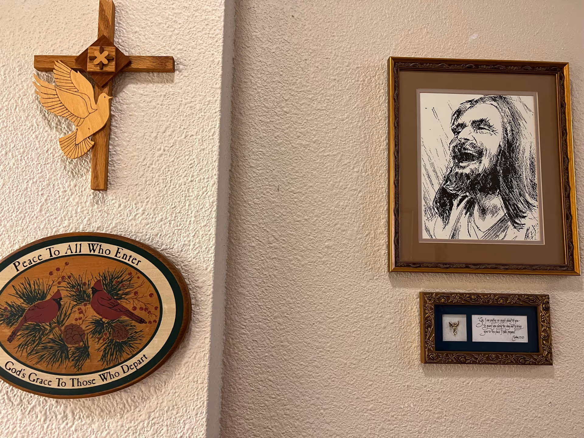 A textured wall with four decorative items: a wooden cross with a carved dove, an oval plaque with two cardinals and the text 'Peace To All Who Enter, God's Grace To Those Who Depart,' a framed black and white sketch of a man with long hair and a beard smiling, and a smaller framed piece with a small angel figure and a handwritten quote.