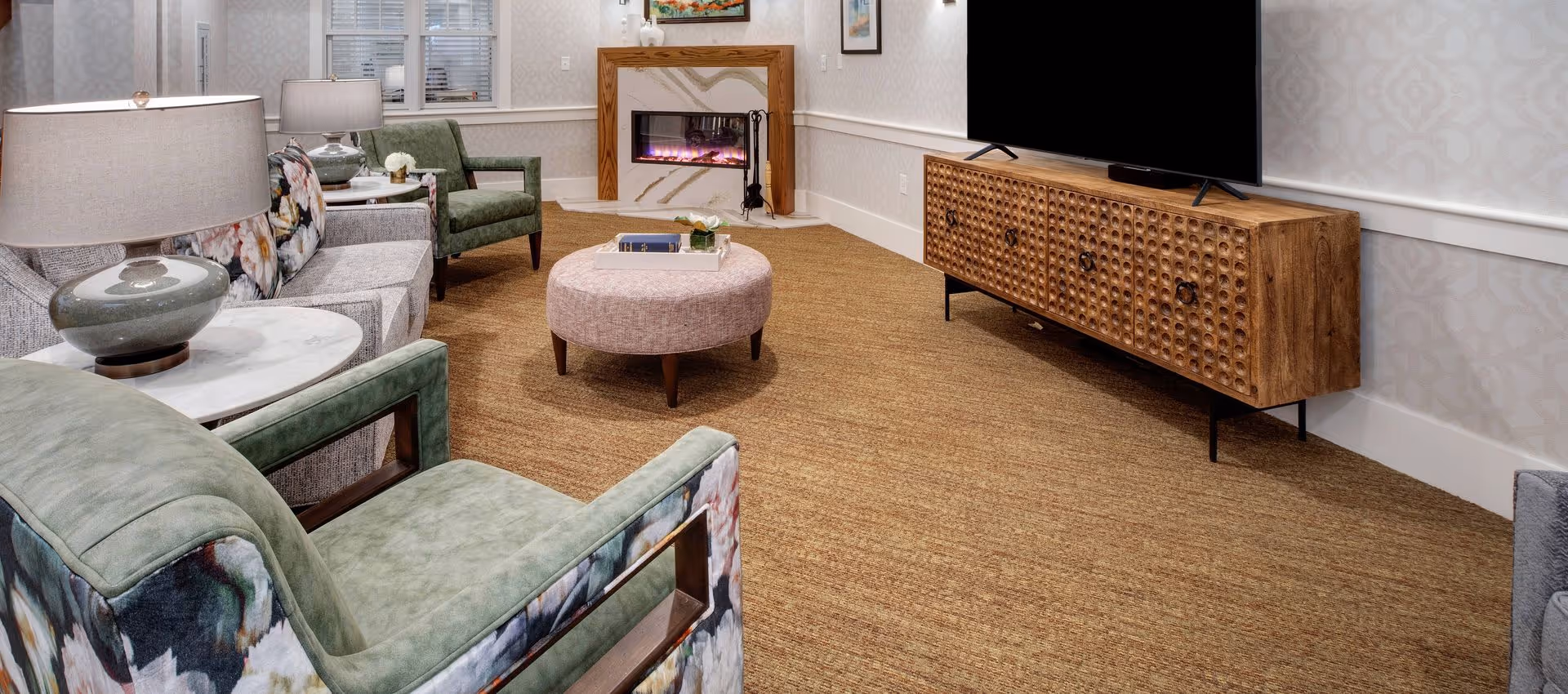 Cozy living room with upholstered chairs, a round ottoman, a TV on a wooden console and an electric fireplace.