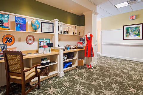 Interior view of a memory care facility hallway with a built-in desk and shelving unit displaying various decorative items, books, and a globe. A wooden chair is placed at the desk. A mannequin dressed in a red and black dress with matching shoes stands near the corner. The carpet has a green and beige floral pattern, and there is a framed painting on the wall near an exit sign.