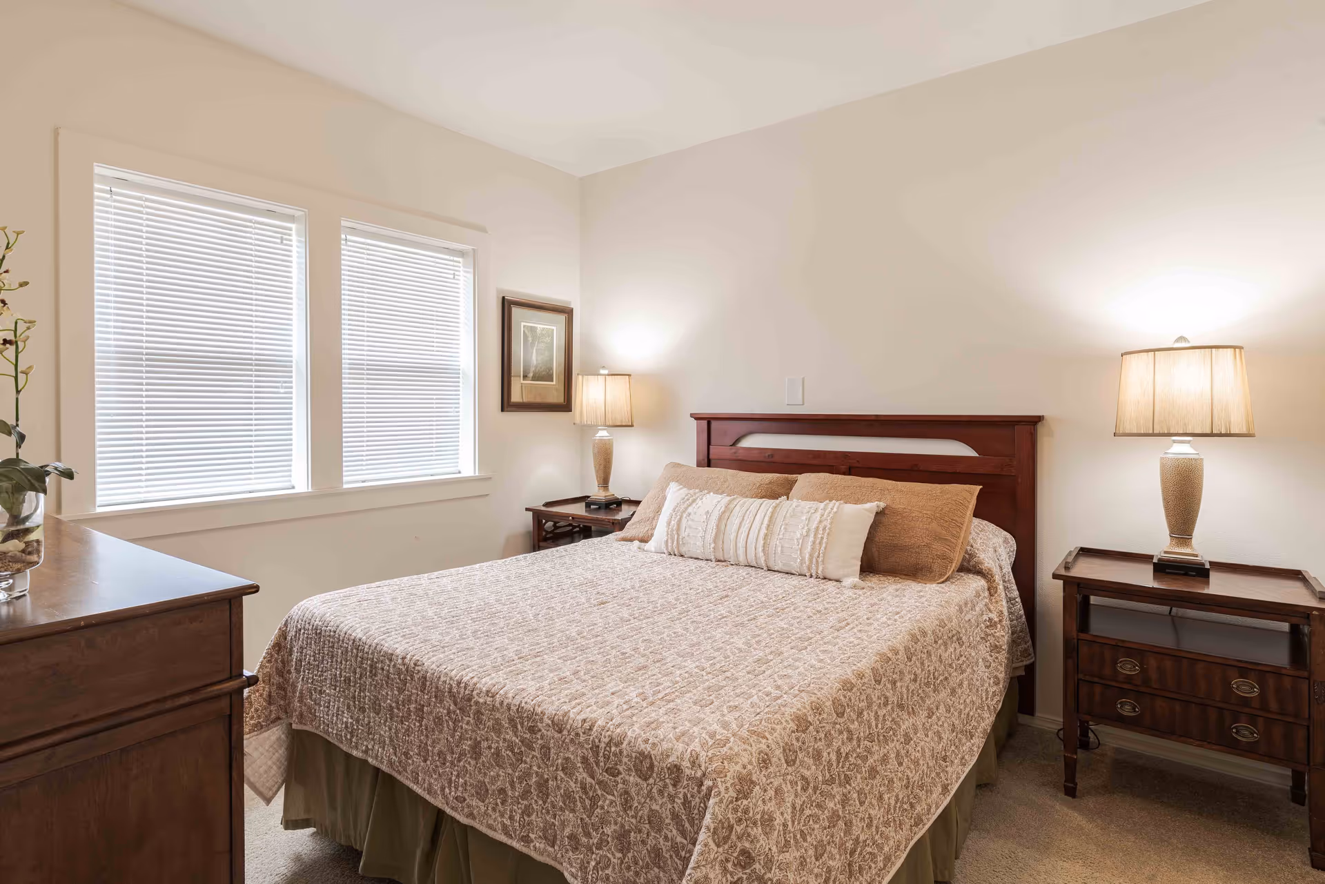 A cozy bedroom with a large bed covered in a beige patterned quilt and decorative pillows. The room has two wooden nightstands, each with a lamp, and a wooden dresser. Two windows with white blinds allow natural light into the room. A framed picture hangs on the wall between the windows and the bed.