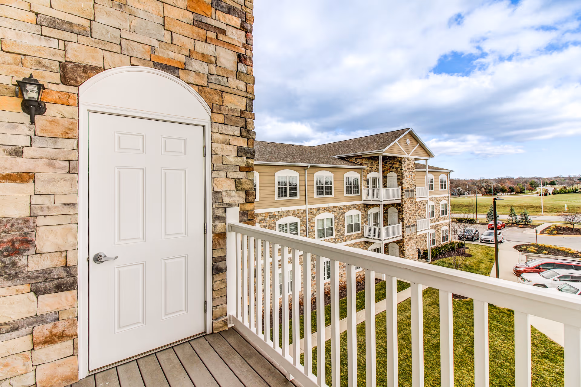 View from a balcony with white railing overlooking a stone and beige siding building with multiple windows and balconies. The scene includes a parking lot with cars and a grassy area under a partly cloudy sky.