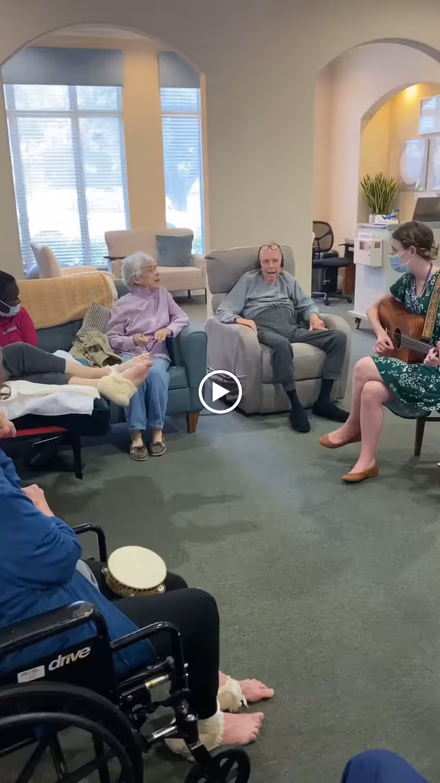 Elderly residents seated in a communal lounge circle while a masked woman plays guitar for them.