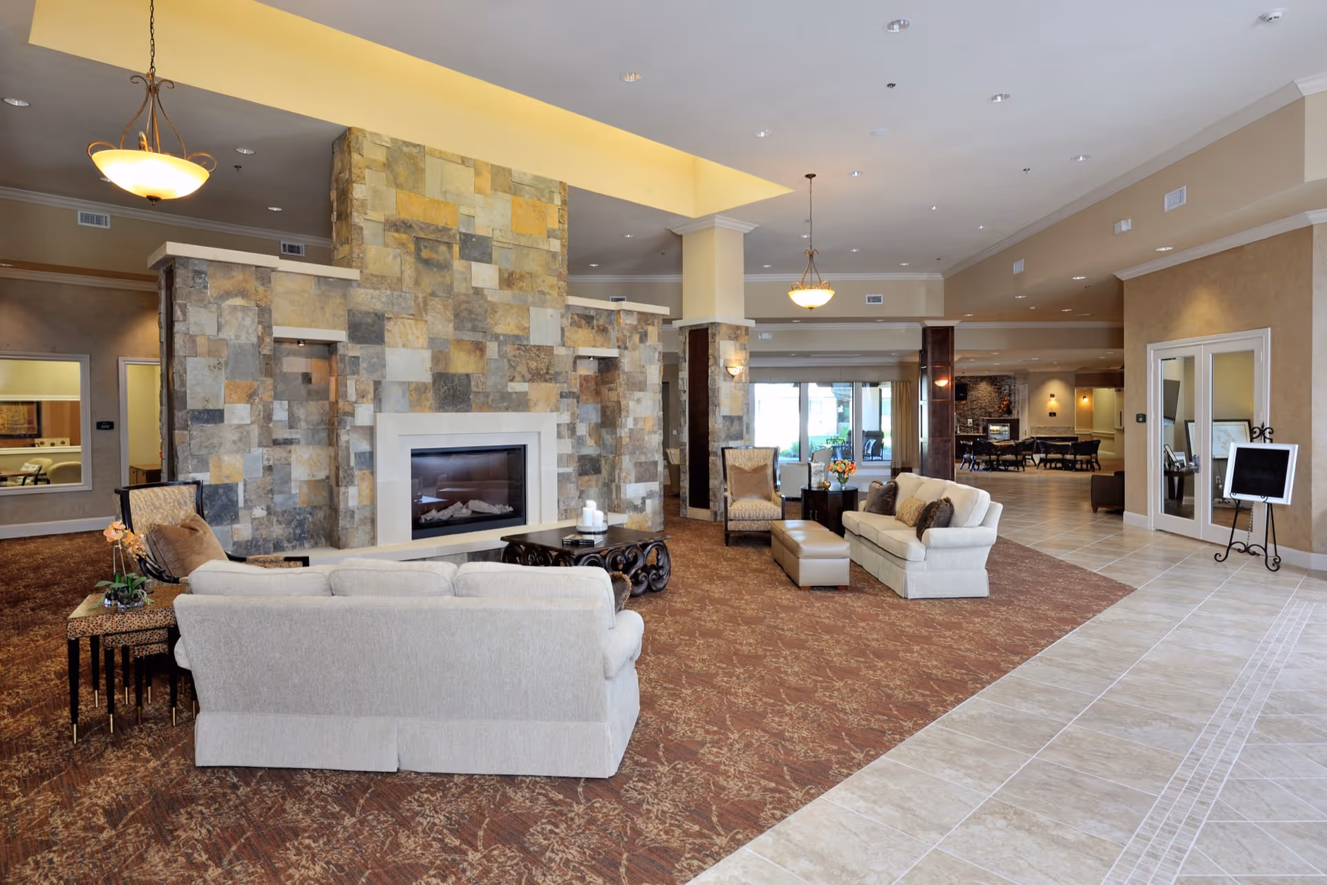 Spacious living room area with beige sofas, armchairs, and a large stone fireplace in the center. The room features a mix of carpet and tile flooring, warm lighting from hanging lamps, and large windows in the background allowing natural light to enter.
