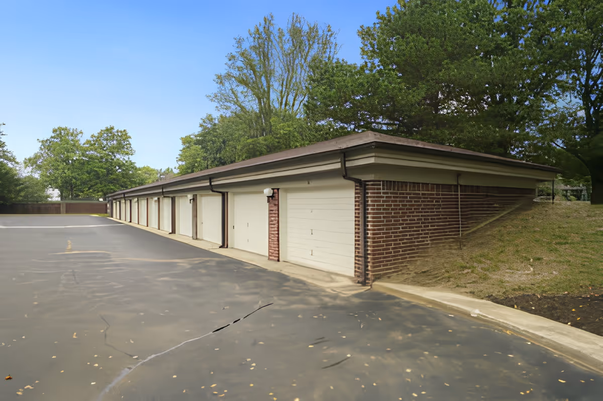 A row of closed garage doors attached to a brick building with a paved driveway in front and trees in the background under a clear blue sky.