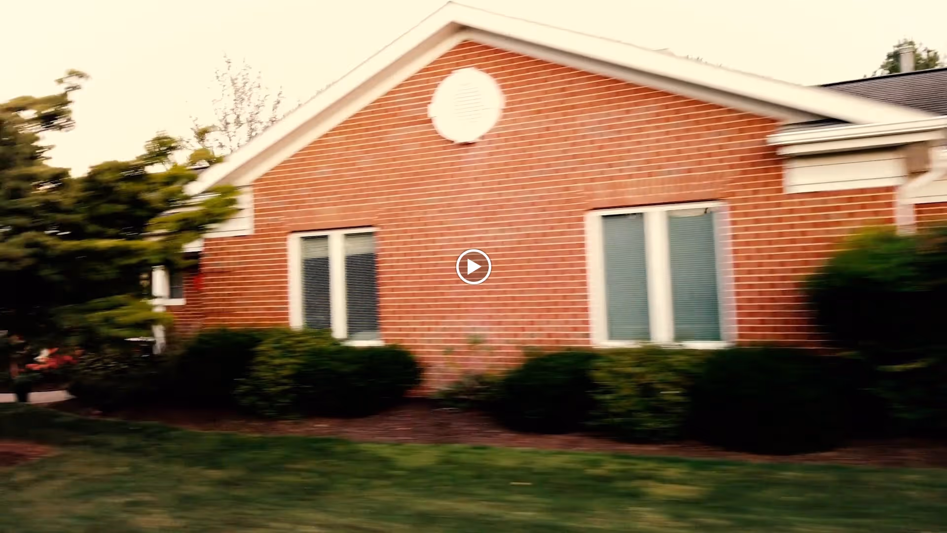 Exterior view of a single-story brick building with white trim, two sets of windows, and bushes along the foundation. There is a grassy lawn in front and trees on the left side.