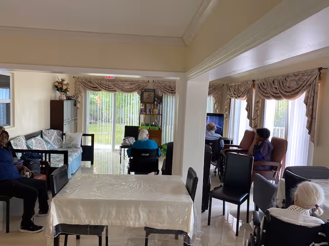 Interior view of a senior living facility common area with several elderly individuals seated in wheelchairs and chairs. The room has large windows with vertical blinds and decorative valances, letting in natural light. There is a table covered with a white tablecloth in the foreground, and comfortable seating arranged around the room.