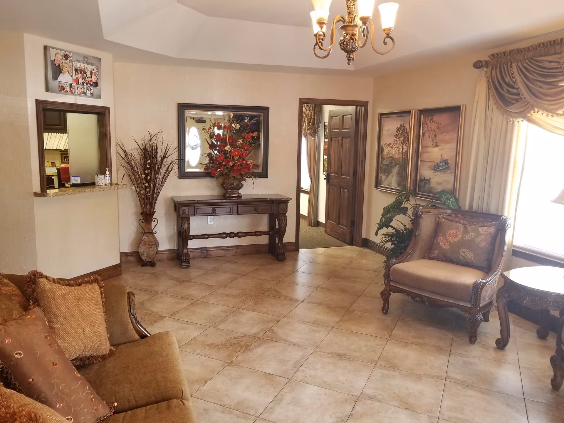 Reception-style sitting area with upholstered chairs and sofa, a console table with floral arrangement, wall art, and a chandelier.