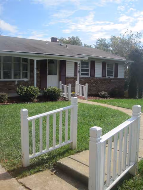 Single-story brick building with white siding, front door, and white picket fence.