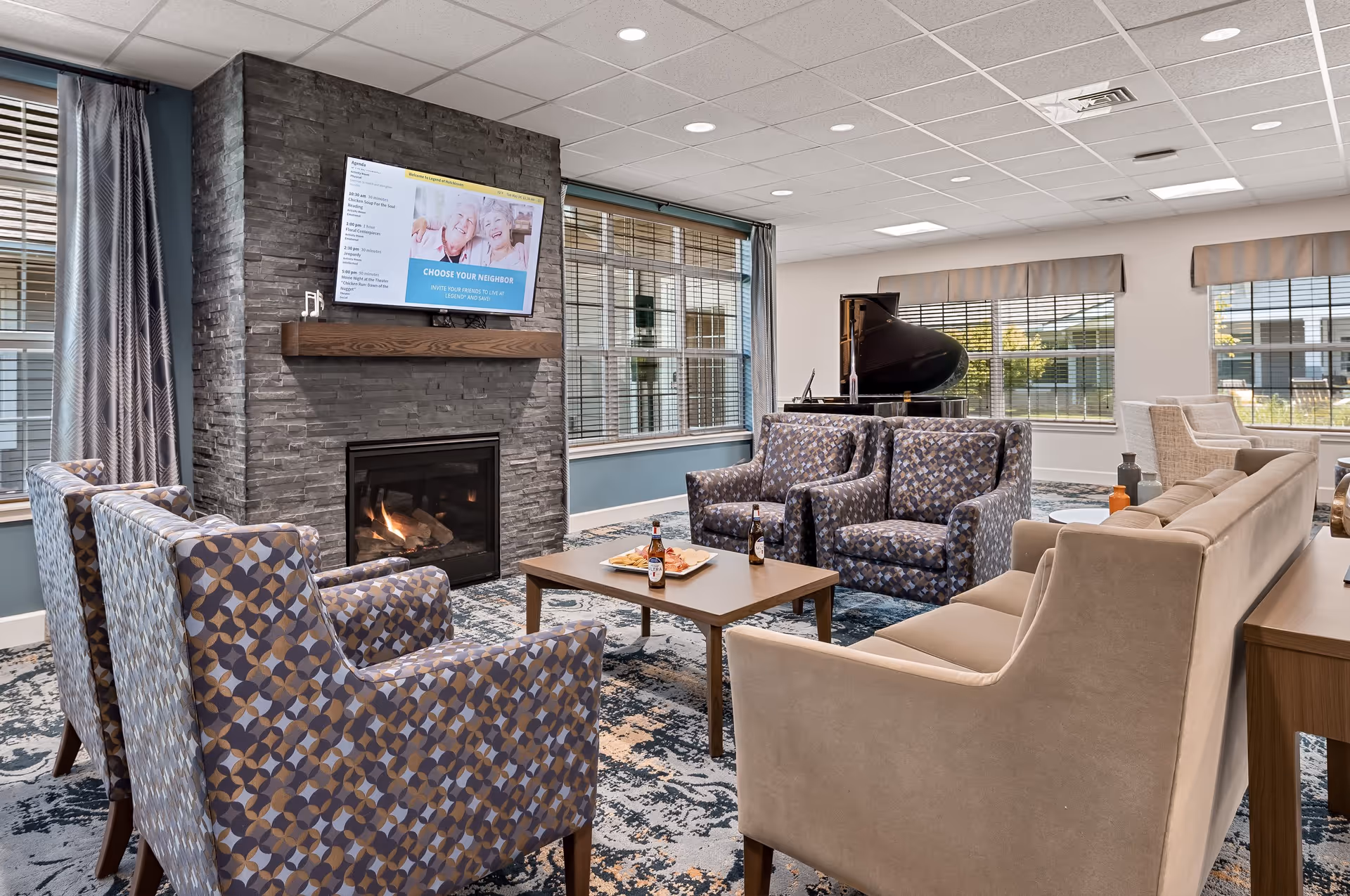 A cozy senior living facility lounge with patterned armchairs, a beige sofa, a wooden coffee table with drinks and snacks, a stone fireplace with a mounted TV above it, large windows with blinds, and a grand piano in the background.