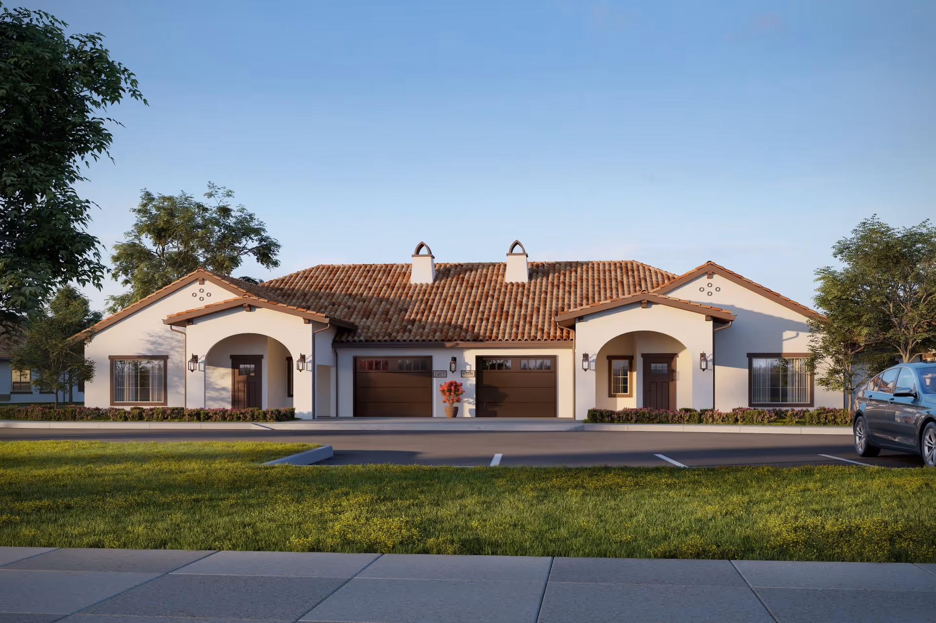 Exterior view of a single-story residential building with a tiled roof, two garage doors in the center, and two front entrances with arched porches on either side. There are trees and shrubs around the building, a parked car on the right, and a clear blue sky above.
