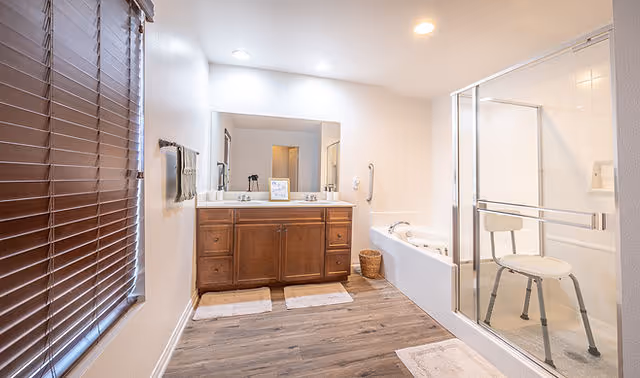 A bright bathroom with wooden flooring, a large mirror above a wooden vanity with two sinks, a bathtub with a grab bar, and a glass-enclosed shower with a white shower chair. There are brown blinds on the window and several white bath mats on the floor.