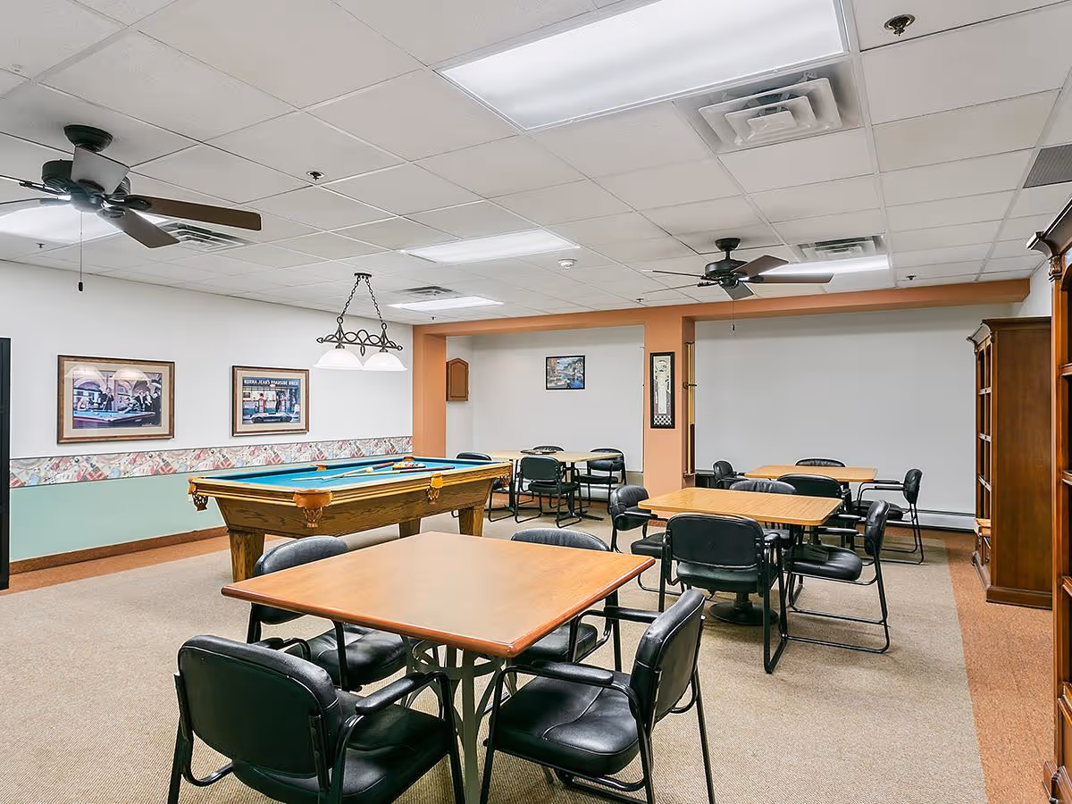 A recreational room with several tables and black chairs arranged for group seating. A pool table with cues and balls is positioned near the wall. The room has ceiling fans, fluorescent lighting, framed pictures on the walls, and a wooden cabinet on the right side.