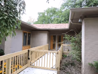 Exterior view of a single-story building with beige brick walls and a wooden ramp leading to a glass door entrance, surrounded by greenery and trees.