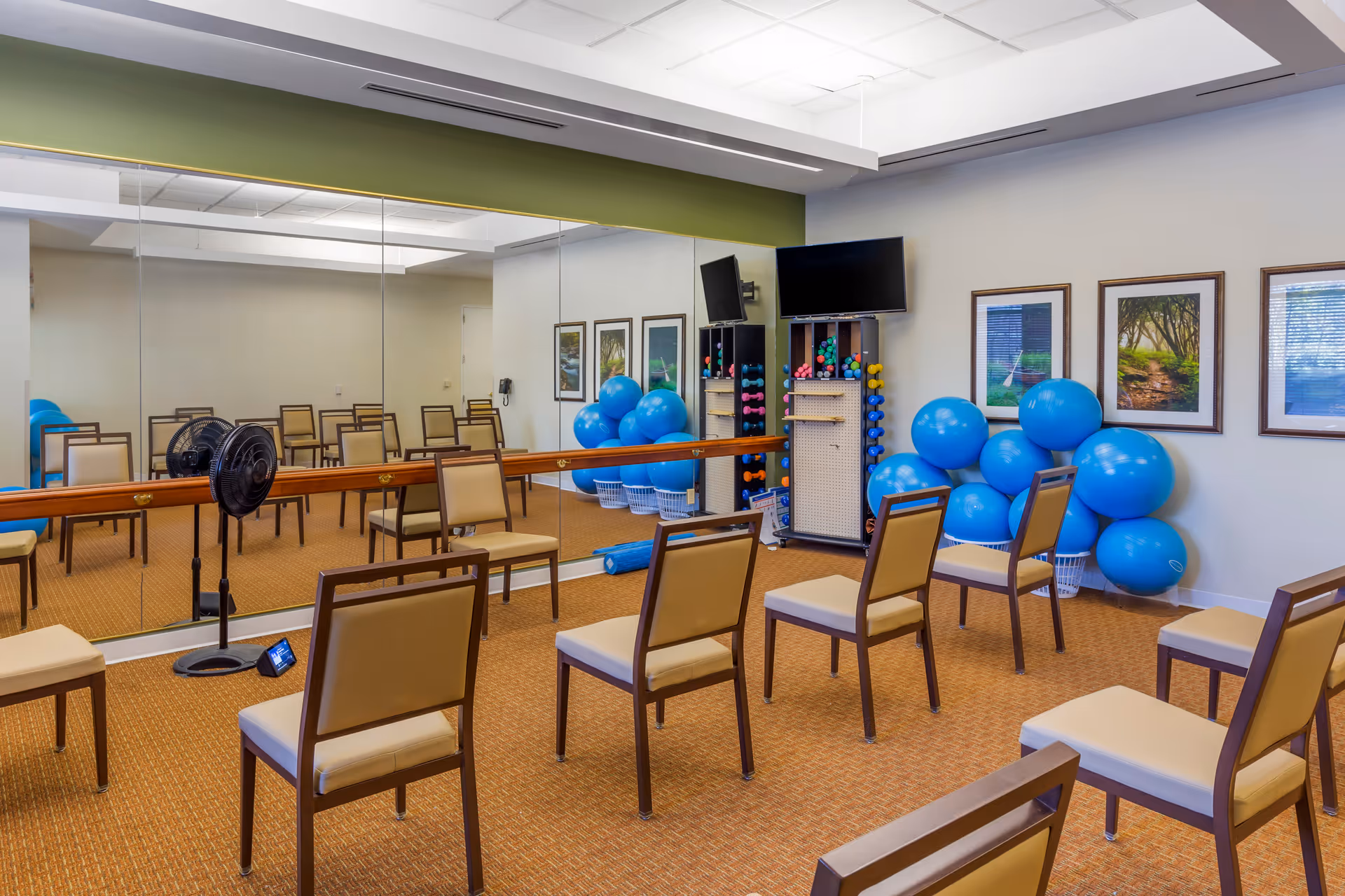A mirrored exercise/activity room with chairs arranged in rows, blue stability balls, hand weights, a wall-mounted TV, and a fan.