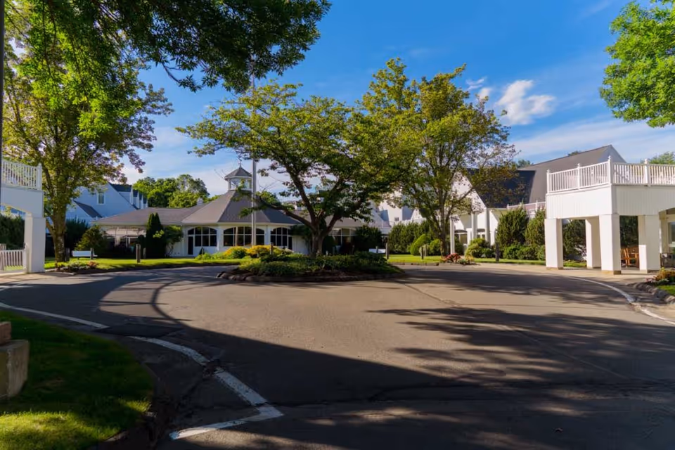 Exterior view of Pomperaug Woods facility showing a circular driveway with a landscaped island in the center, surrounded by white buildings with pitched roofs and large windows, under a clear blue sky with trees providing shade.