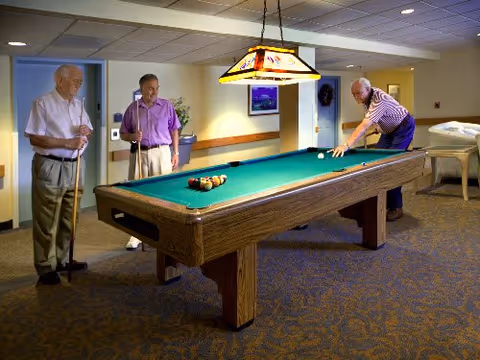 Three elderly men playing pool in a well-lit room with a green pool table, carpeted floor, and a hanging light fixture above the table. Two men stand on one side holding pool cues, while the third man is bent over aiming a shot. The room has beige walls and some furniture in the background.