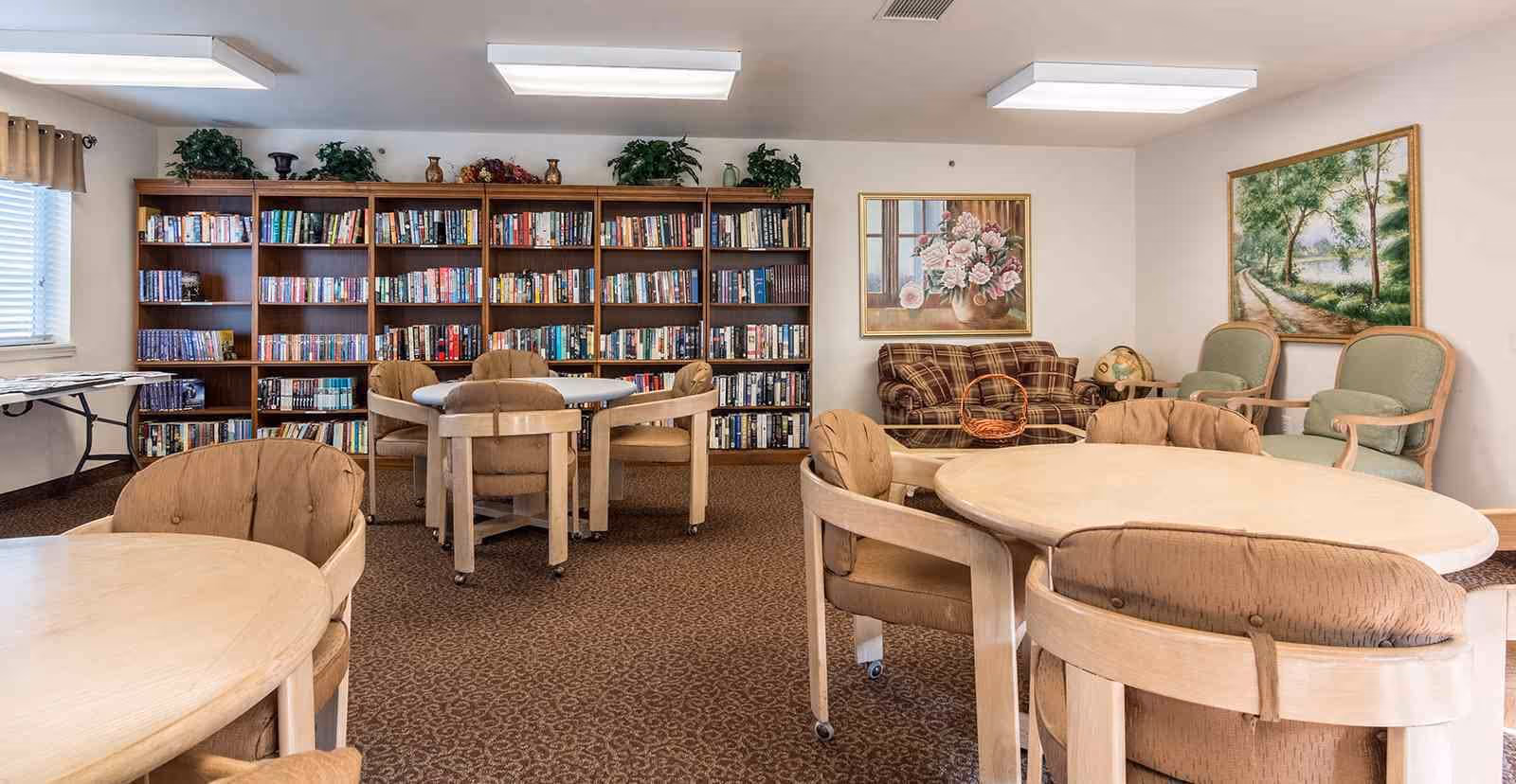 A cozy common room with round tables and cushioned chairs, a large bookshelf filled with books, a plaid sofa, two green armchairs, and framed paintings on the walls. The room has carpeted flooring and fluorescent ceiling lights.