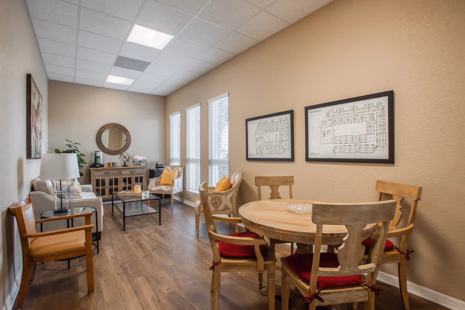 A cozy interior room with wooden flooring, featuring a round wooden table with four chairs that have red cushions. On the right wall, there are two framed floor plans. The left side of the room has a white sofa, a wooden armchair, a glass-top coffee table, and two patterned armchairs with yellow cushions. A wooden cabinet with a round mirror above it and a coffee maker is positioned against the far wall. Three tall windows with blinds allow natural light into the room.