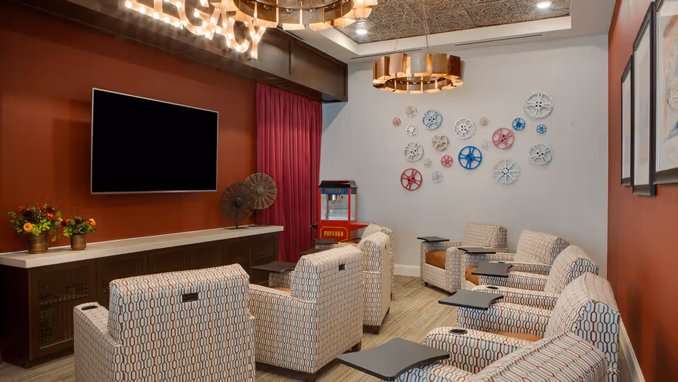 A cozy media room with patterned armchairs arranged in rows facing a large flat-screen TV mounted on a red wall. There is a popcorn machine in the corner and decorative film reels mounted on a white wall. The ceiling has a decorative design with warm lighting fixtures, and a sign above the TV reads 'LEGACY'.