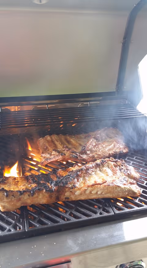 Two racks of ribs cooking over open flames on a grill with smoke rising.
