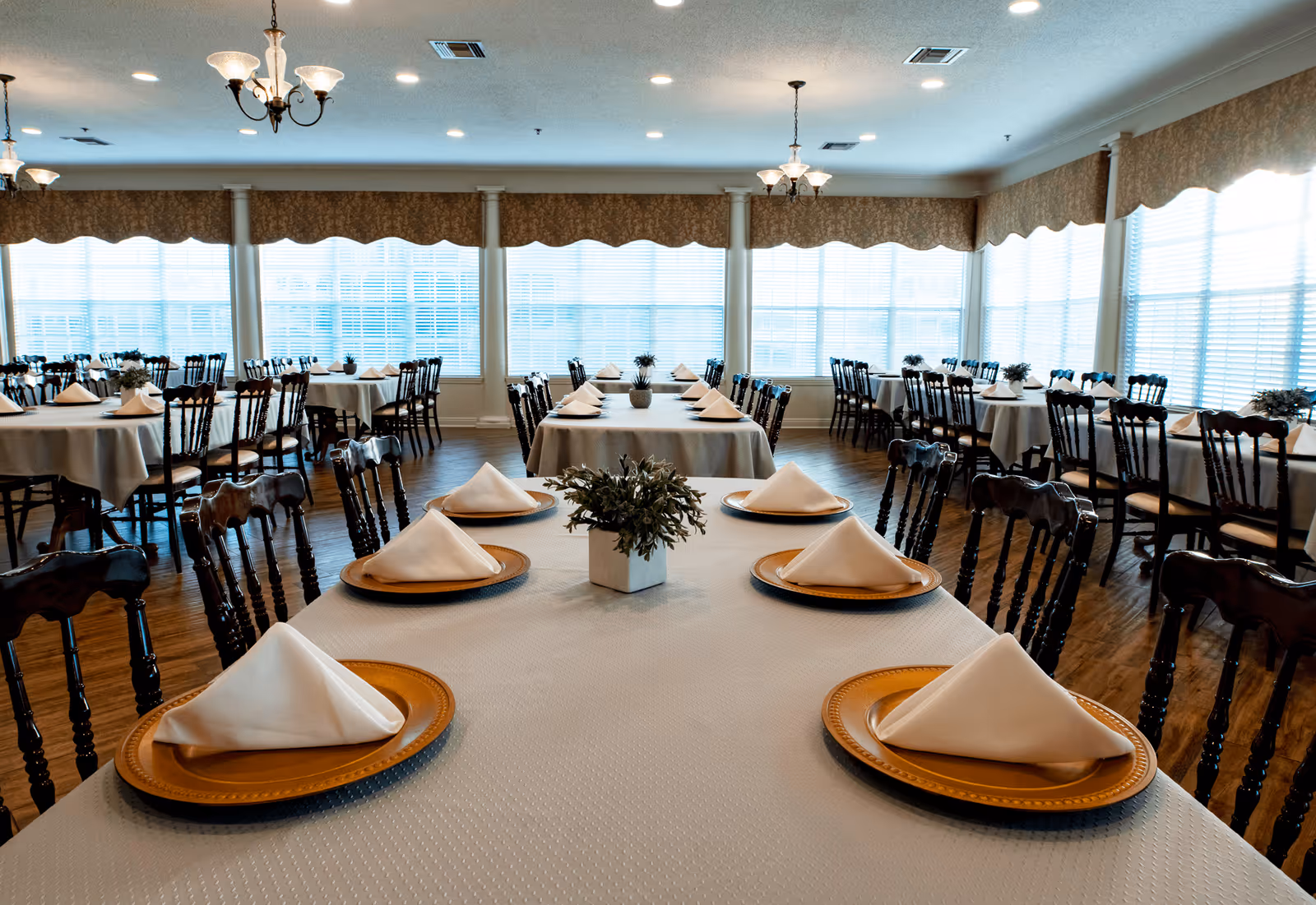 A bright dining room with multiple tables covered in white tablecloths, each set with gold charger plates and neatly folded white napkins. The room features wooden chairs, large windows with blinds, and decorative valances. Small potted plants serve as centerpieces on the tables, and elegant chandeliers hang from the ceiling.