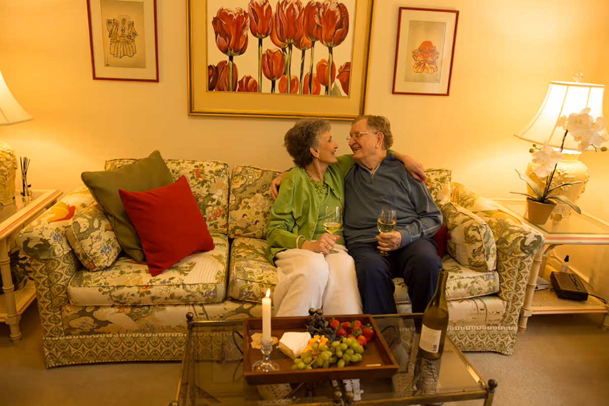 An elderly couple sitting closely on a floral patterned couch in a warmly lit living room, holding glasses of white wine and smiling at each other. A glass coffee table in front of them holds a tray with grapes, strawberries, cheese, and a lit candle. The room is decorated with framed floral artwork on the wall and table lamps on side tables.