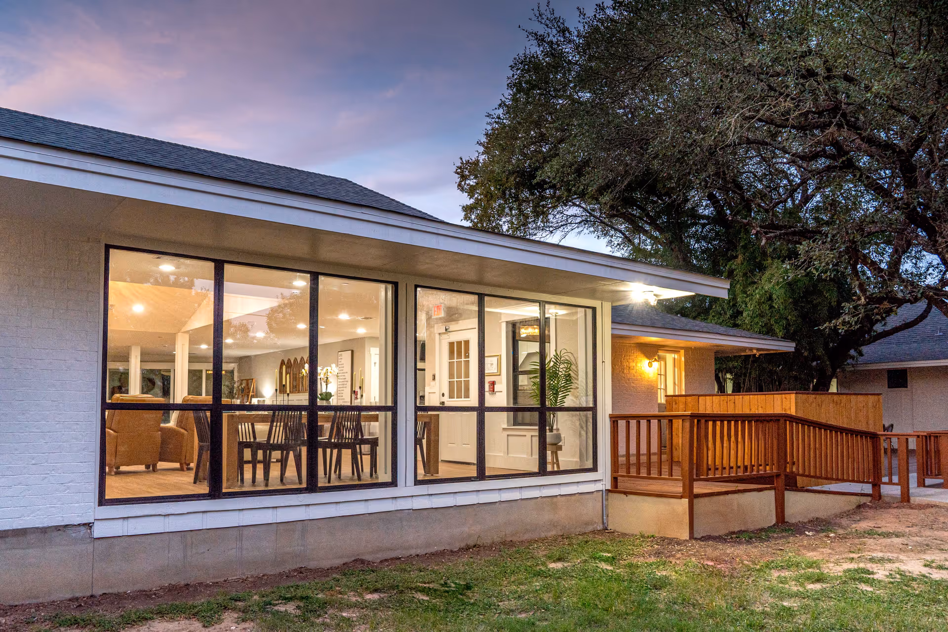 Exterior view of a single-story assisted living facility building at dusk, showing large windows revealing a well-lit interior with dining tables and chairs. There is a wooden ramp and railing leading to the entrance, surrounded by grass and large trees.