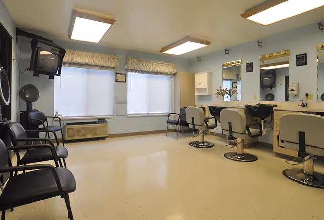 Interior of a hair salon area in a senior living facility with several salon chairs facing mirrors, a hair dryer, and waiting chairs along the opposite wall. Two large windows with blinds and valances provide natural light.