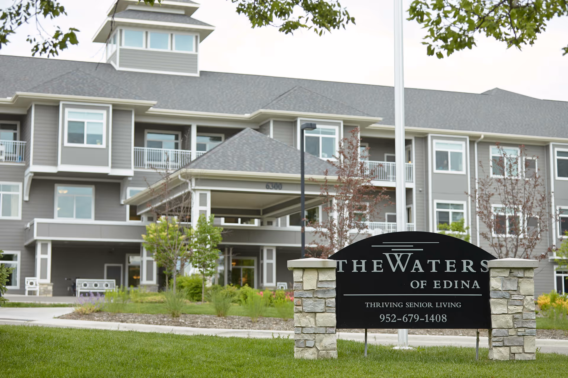 Front exterior of The Waters of Edina senior living building with an entrance canopy and a stone sign on the lawn.