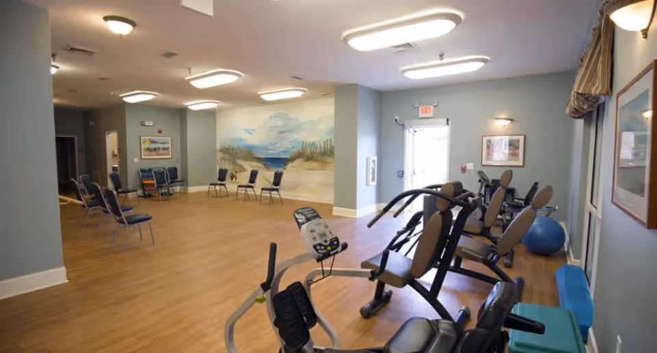 A spacious exercise room with several pieces of fitness equipment including stationary bikes and weight machines. The room has wooden flooring, light blue walls, and ceiling lights. There are several chairs arranged along the far wall, which features a mural of a beach scene. A door with an exit sign is visible at the back.