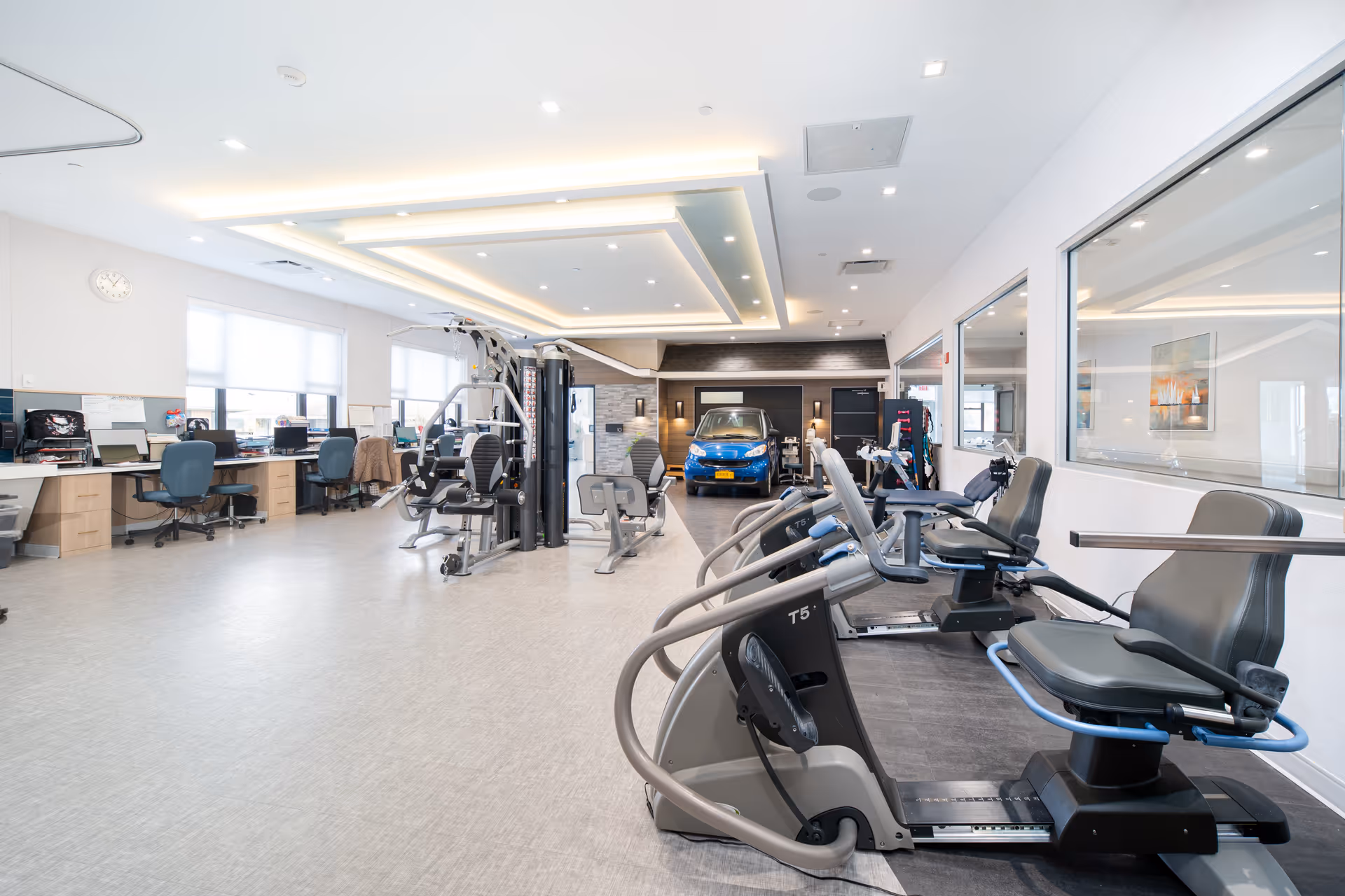 A spacious, well-lit rehabilitation gym area with exercise equipment including recumbent bikes and strength training machines. There are desks and chairs along the left wall with computers, and a blue car is parked at the far end of the room near a dark accent wall. Large windows and ceiling lights provide ample lighting.