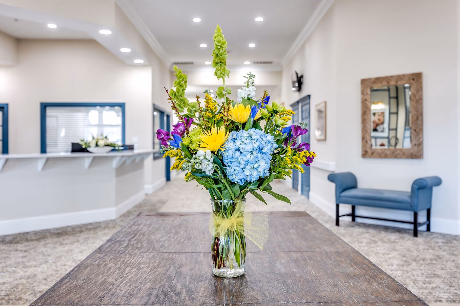 A bright and clean interior space with a wooden table in the foreground holding a glass vase filled with a colorful bouquet of flowers including blue hydrangeas, yellow daisies, and purple and green blooms. The background shows a reception desk with a flower arrangement, a blue cushioned bench, a large mirror on the wall, and a hallway with recessed lighting.