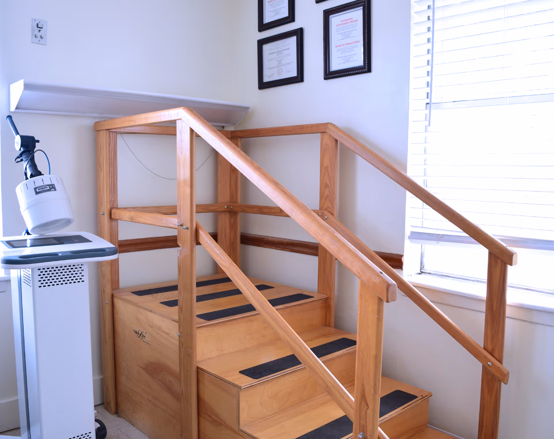 A small wooden rehabilitation staircase with handrails on both sides, placed indoors near a window with blinds. Next to the staircase is a medical device with a mounted light. Three framed certificates or documents are hanging on the wall above the staircase.