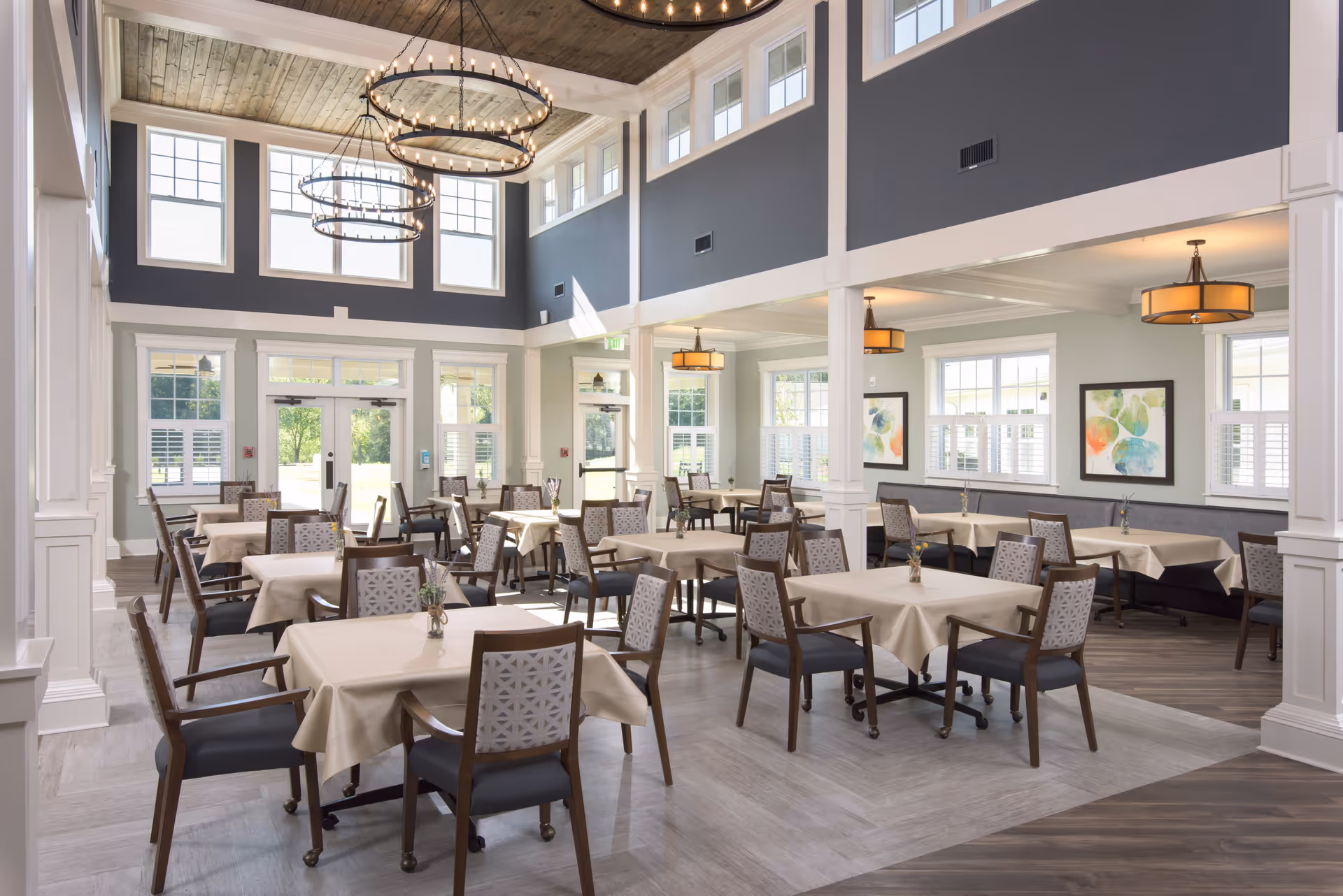A bright and spacious dining room with multiple tables covered in beige tablecloths, each surrounded by wooden chairs with patterned backs. The room features large windows allowing natural light to fill the space, high ceilings with wooden accents, and modern circular chandeliers. There are framed abstract paintings on the walls and a mix of light and dark flooring.