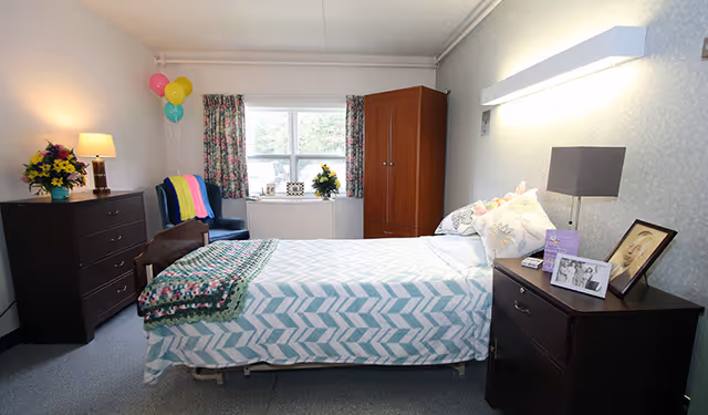 Well-lit single bedroom with a made bed, dark wood dresser and nightstand, wardrobe, armchair by the window, and floral decorations.