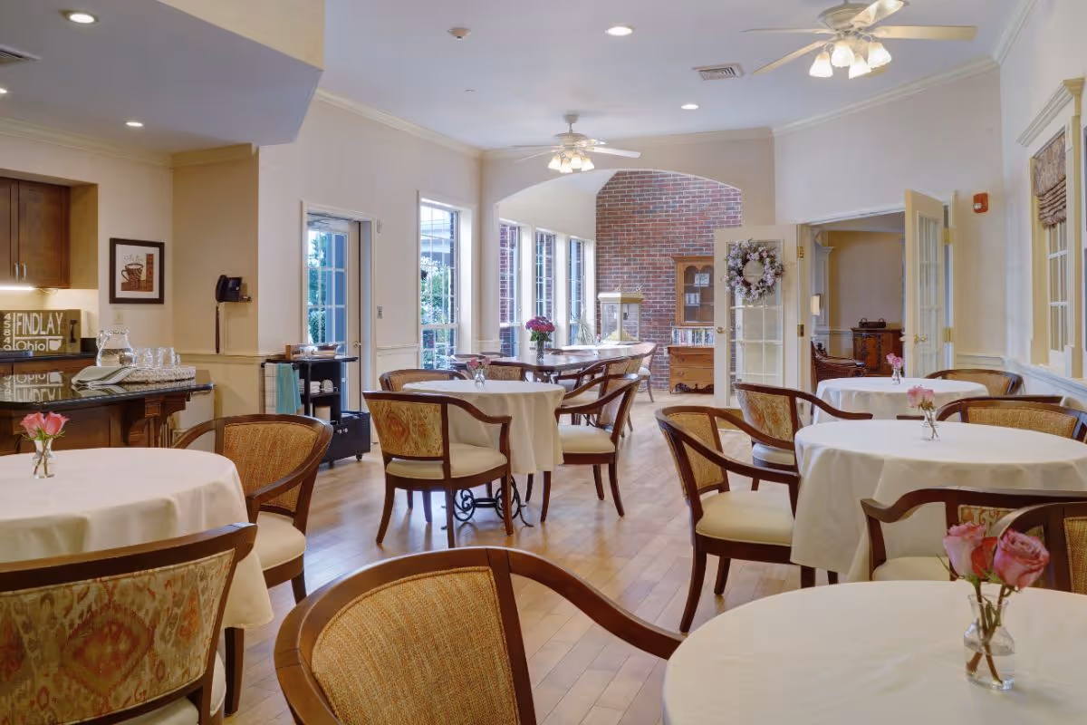Bright dining room with round tables draped in white tablecloths, wooden chairs, small vases of flowers, and a serving area near windows.