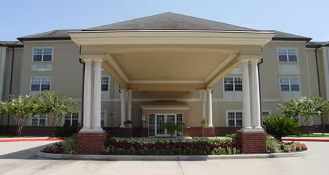 Front exterior view of a multi-story senior living facility with a large covered entrance supported by white columns, landscaped greenery, and a clear sky above.