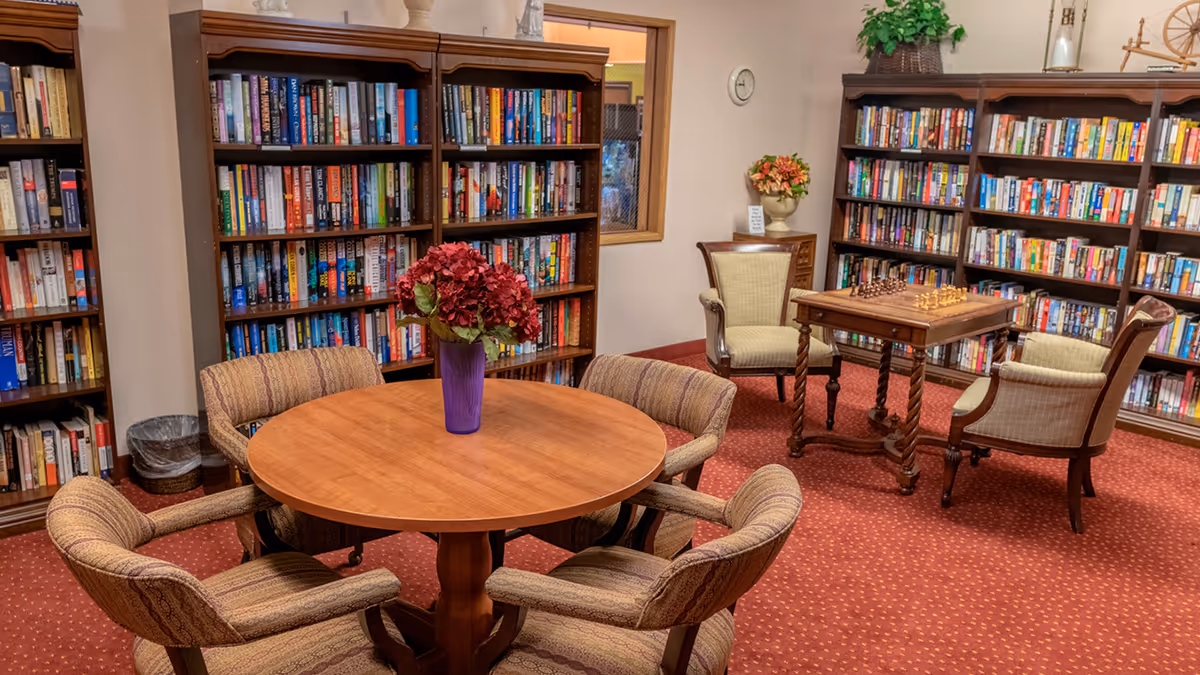 A cozy library room with wooden bookshelves filled with books along the walls. In the center, there is a round wooden table with a purple vase holding red flowers, surrounded by four upholstered chairs. To the right, a small wooden table with a chessboard is flanked by two armchairs. The room has a red carpet and soft lighting.