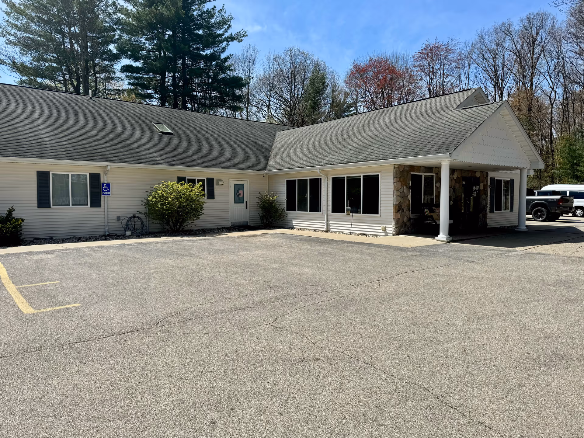 Exterior view of a single-story building with white siding and a gray roof, surrounded by trees. The building has large windows and a covered entrance supported by white columns. There is a parking lot in front with a designated handicapped parking space.