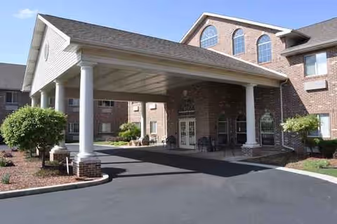 Entrance of a senior living facility with a covered driveway supported by white columns, brick exterior walls, and landscaped areas with bushes and mulch.