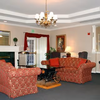 Communal lounge with patterned red sofas and armchairs around a coffee table, chandelier, fireplace, and framed artwork.