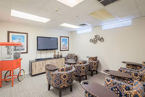 A small media room with patterned armchairs arranged in rows facing a wall-mounted flat screen TV. There is a wooden cabinet beneath the TV with decorative items on top. On the left side of the room, there is a red popcorn machine. The walls are light-colored with two framed pictures and a decorative metal wall art piece. The ceiling has recessed lighting and air vents.